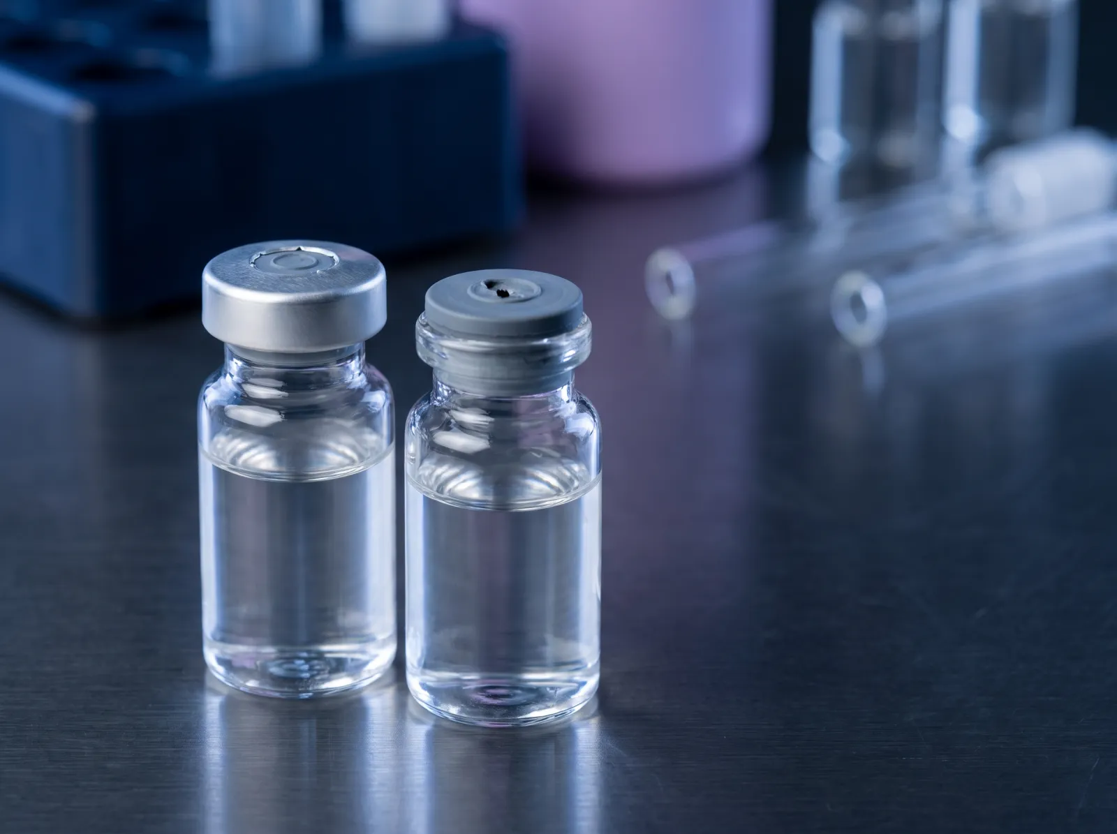 Sealed unopened vial beside an opened vial with a punctured grey stopper on a laboratory bench, close up