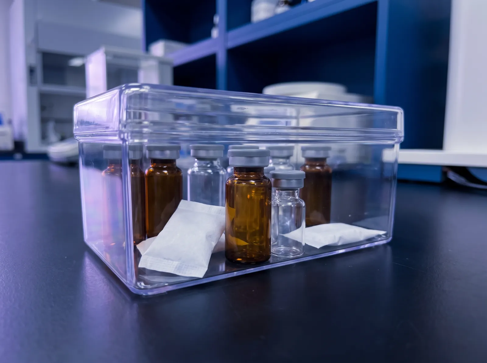 Amber and clear glass vials with desiccant packets in a sealed clear container on a dark laboratory bench