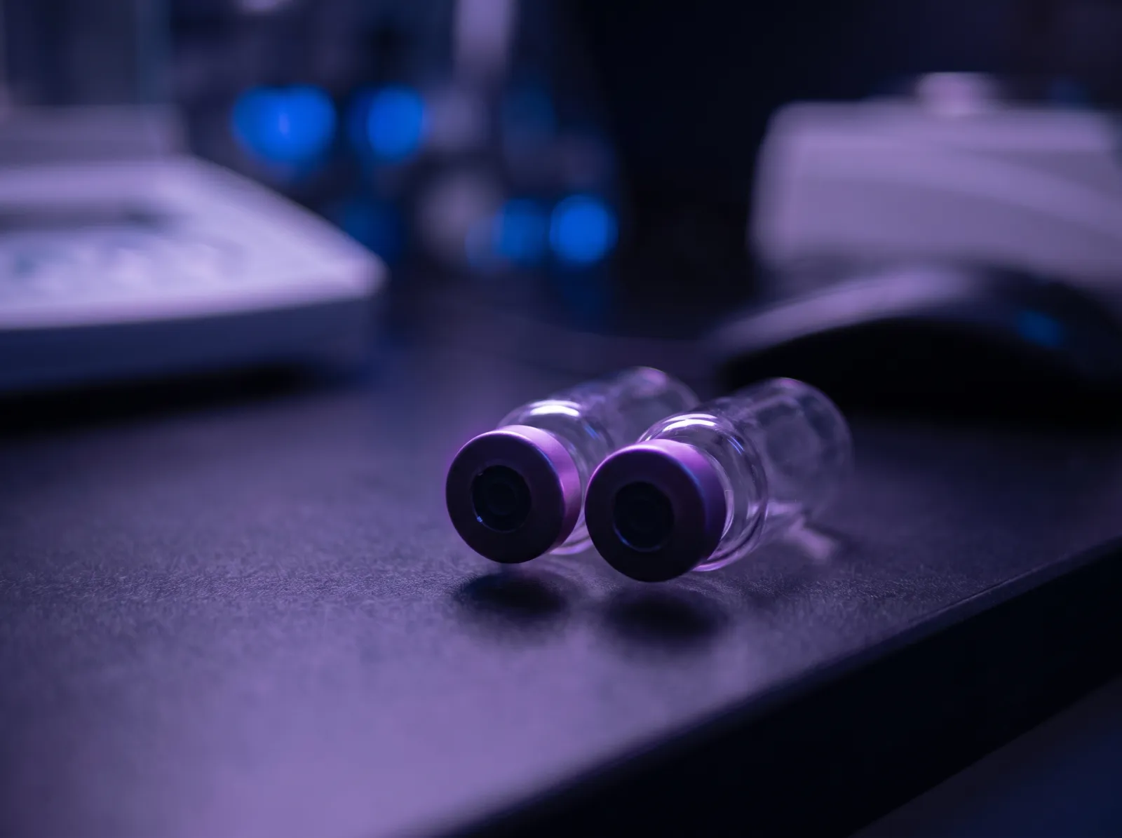 Two small unlabeled glass vials side by side on a dark laboratory bench with soft violet rim light